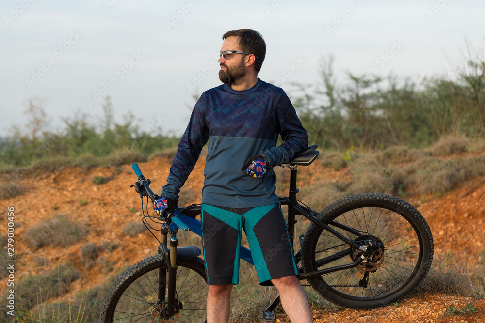 Fototapeta premium Cyclist in shorts and jersey on a modern carbon hardtail bike with an air suspension fork rides off-road on the orange-red hills at sunset evening in summer 