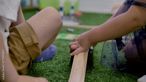Two children, a boy and a girl, playing on a plastic toy, sitting on the green artificial grass in the room.