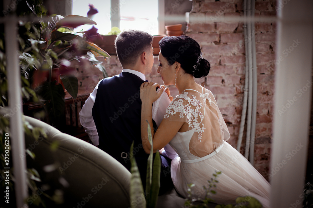 Naklejka premium Groom in vest and beautiful bride in an elegant dress sitting back on the sofa among the houseplants in a stylish interior