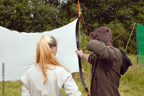 A girl in folk costume is watching a man in modern clothes shooting a backpack from a medieval bow. Camping on the weekends at a historic festival. Training in shooting from the national weapon. photo