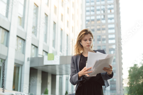 Concentrated stylish businesswoman standing on city street with documents reading them. Successful female manager dressed in elegant suit checking accounting on paper during coffee break outdoors.