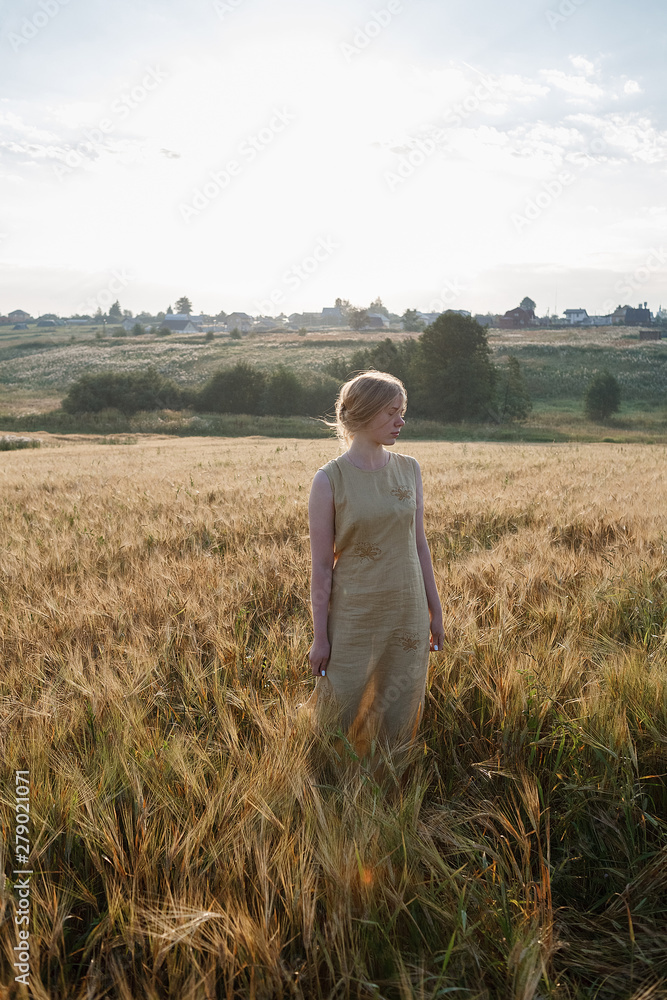 young pretty girl in yellow dress stands at field of ears in rays of rising sun. grove and village in background young pretty girl in yellow dress stands at field of ears in rays of rising sun. grove and village in background