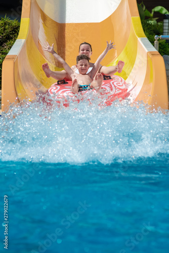 Caucasian boy and mom gliding down slide in waterpark. They enjoy the fun and holding hands wide open.