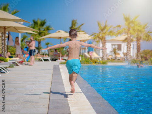 Caucasian boy having fun in swimming pool at resort. He is running along swimming pool. His arms are wide open.