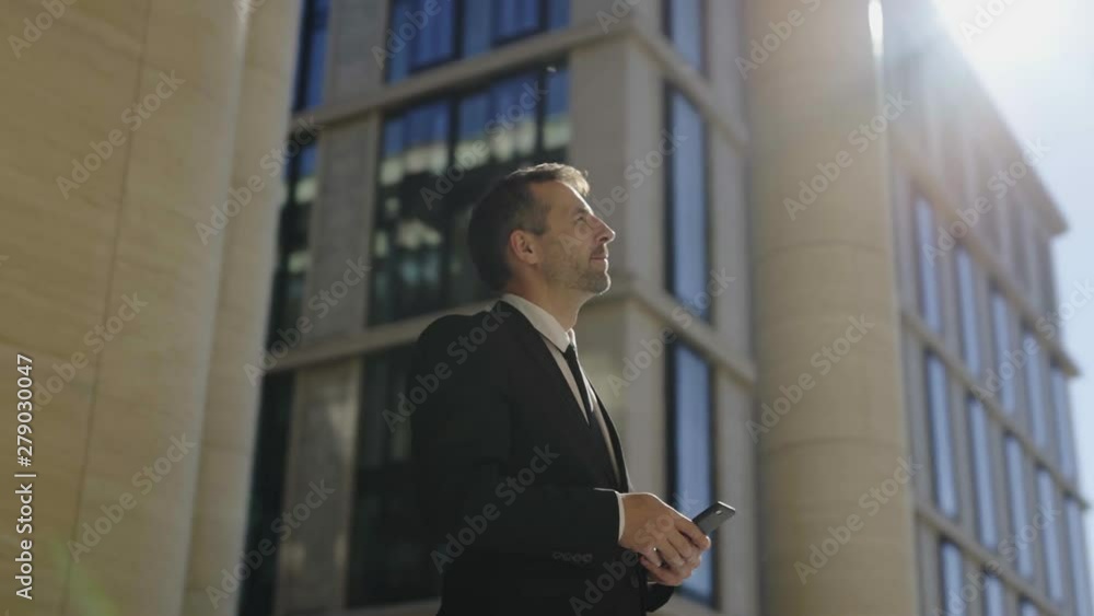 Panning waist up shot of middle aged businessman in black formal suit and necktie standing outside and browsing cell phone. Man looking up and thinking in sunlight