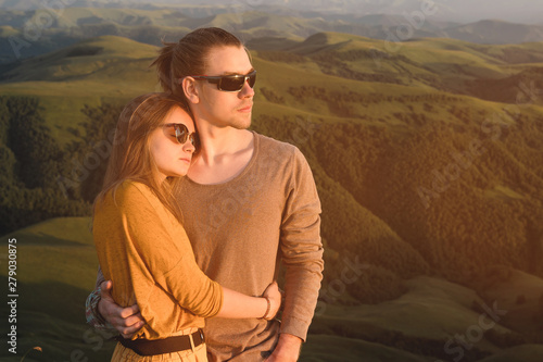 Portrait of a romantic young hipster couple hugging. They stand in an embrace in nature high in the mountains against the backdrop of a mountain green valley and clouds.
