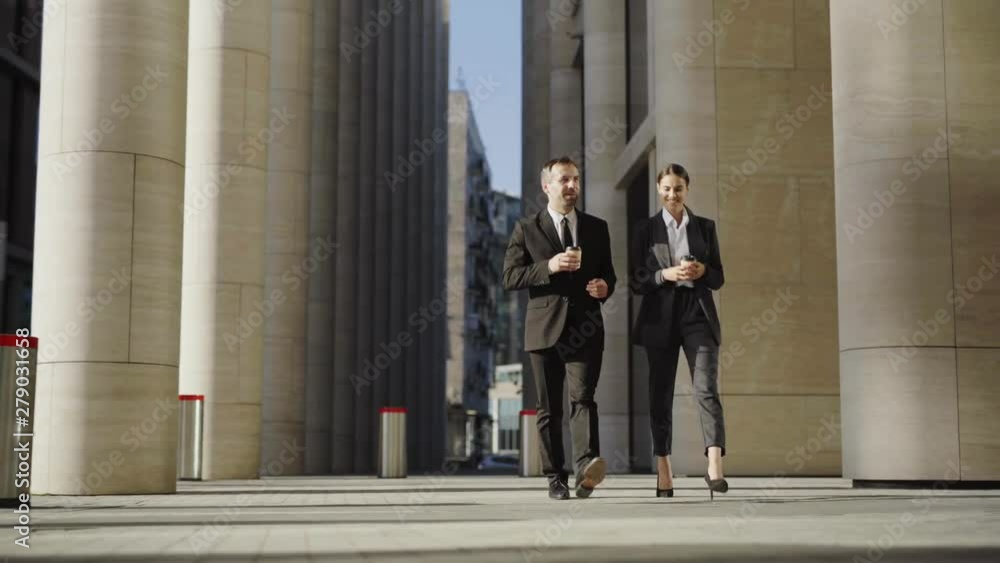 Front view of two business partners, handsome middle aged man and beautiful young woman in black formal suits walking down street together, drinking coffee and talking Front view of two business partners, handsome middle aged man and beautiful young woman in black formal suits walking down street together, drinking coffee and talking