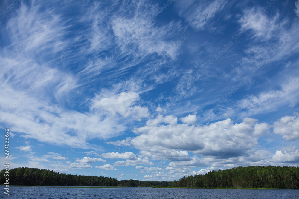 Background with thin white clouds in the blue summer sky over the lake. Forest on the far bank. With copy space