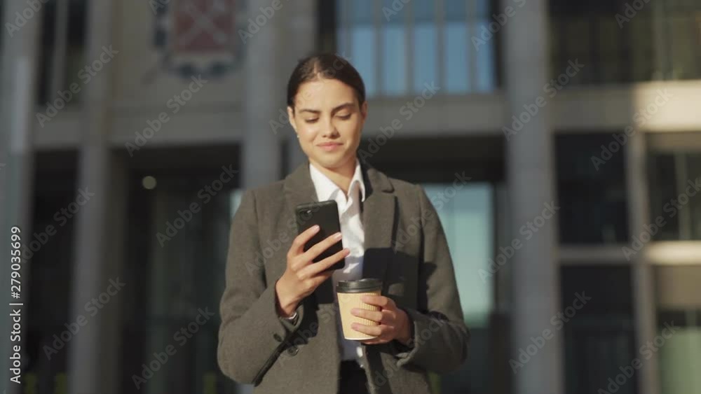 Waist up shot of smiling young businesswoman in coat walking down street, texting on cell phone, looking up dreamily thinking of response and drinking takeaway coffee Waist up shot of smiling young businesswoman in coat walking down street, texting on cell phone, looking up dreamily thinking of response and drinking takeaway coffee