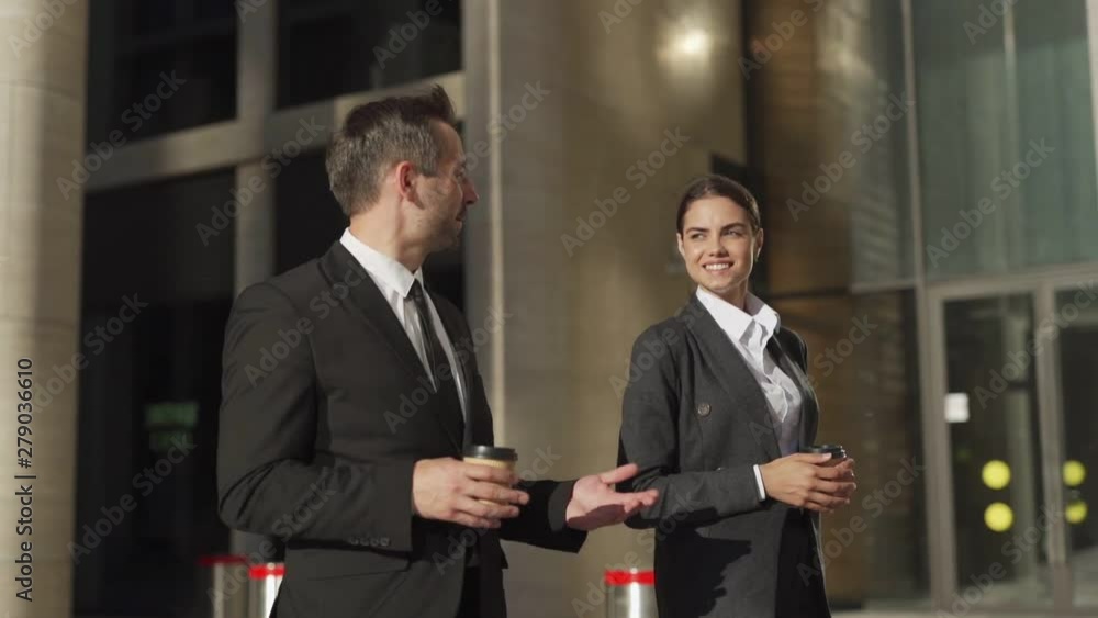 Tracking slow motion shot of two businesspeople in formal suits, middle aged man and young woman walking down street with takeaway coffee cups and talking friendly Tracking slow motion shot of two businesspeople in formal suits, middle aged man and young woman walking down street with takeaway coffee cups and talking friendly