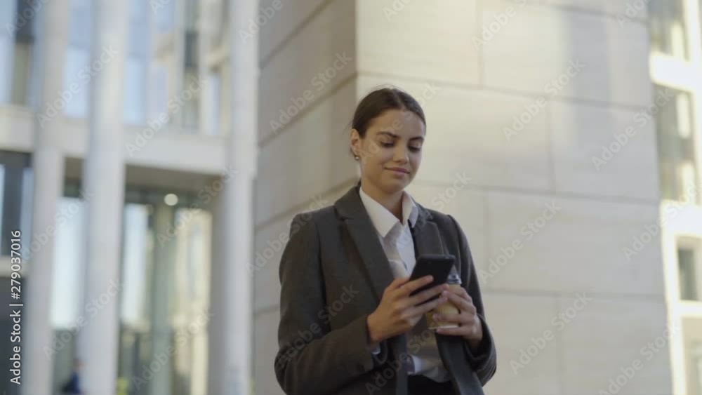 Medium slow motion shot of beautiful young businesswoman in coat walking down city street after work. Woman reading messages on cell phone, drinking takeaway coffee and smiling slightly Medium slow motion shot of beautiful young businesswoman in coat walking down city street after work. Woman reading messages on cell phone, drinking takeaway coffee and smiling slightly