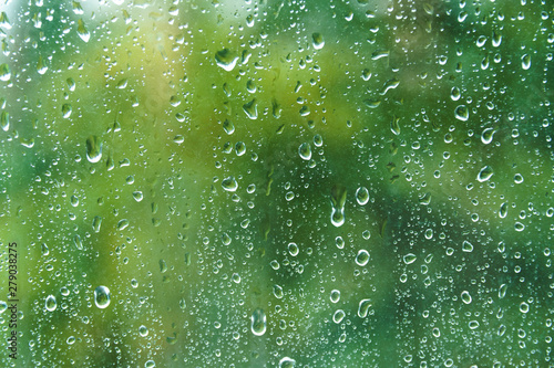 Close up of fresh water drops on window glass on background of green leaves. Raindrops on windowpane in summer day. Droplets on glass with blurry summer garden background.