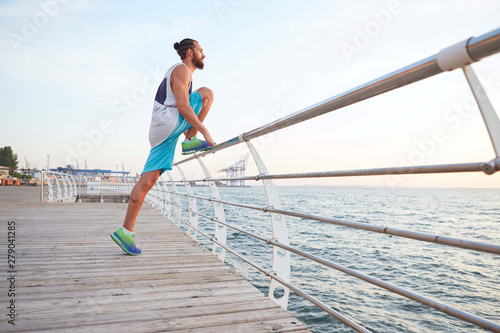 Portrait of young attractive bearded guy doing stretching for legs, morning exercises by the sea, warm-up after run, leads healthy active lifestyle. Fitness male model.