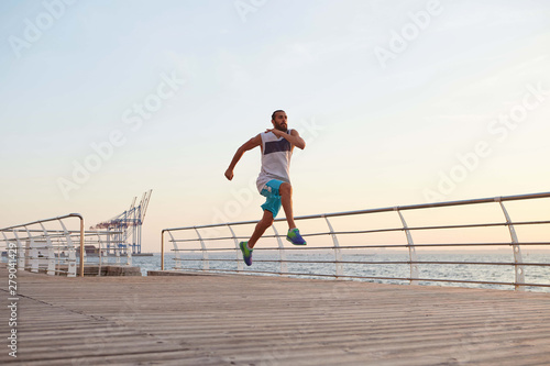 Photo of young sporty handsome bearded running guy doing morning exercises by the sea, warm-up before run, leads healthy active lifestyle. Fitness and sport concept.