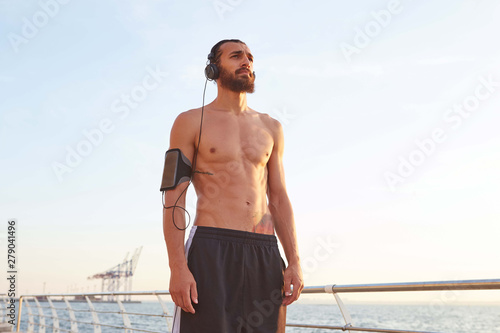 Young attractive sporty bearded man after extreme sport at the seaside, rest after jogging, looking at the sea and listening songs on headphones, leads healthy active lifestyle. Fitness male model.
