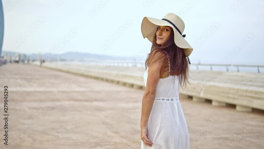 Pretty woman wearing white dress and pamela hat walking over the beach terrace at sea. Happy summer vacation Pretty woman wearing white dress and pamela hat walking over the beach terrace at sea. Happy summer vacation