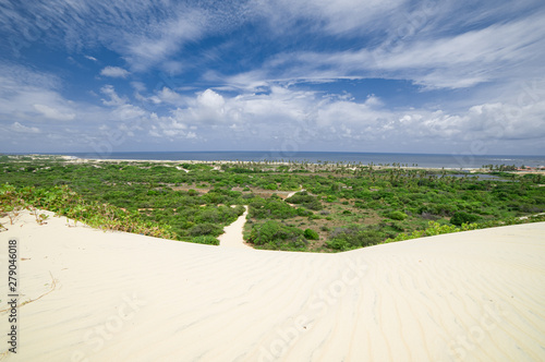 Sand dune leading to a road through a green field and the sea. Cloudy sky. Natal, Rio Grande do Norte, Brazil. photo