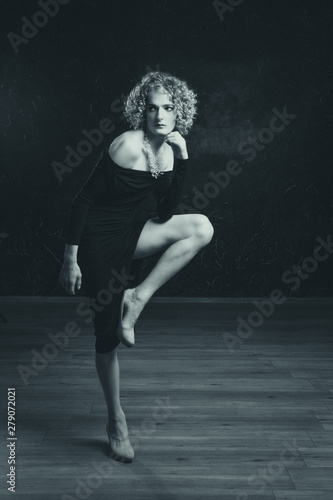 Black and white portrait of a sad transgender guy model with blue eyes and blond hair in the image of a woman an afro hair,standing in an extravagant pose on one leg like a heron.