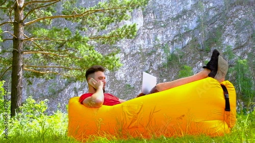 The guy in the red T-shirt lies on the air chair, talking on the phone and working on a laptop. A man with a beard is a freelancer or a gamer. Concept of leisure and business, study and lifestyle. photo