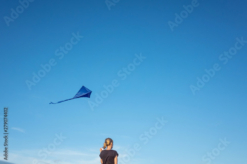 A young girl stands with her back to the frame. She launched a blue kite into a cloudless sky. The concept of freedom, summer hobbies, entertainment in nature. Minimalism, a place for text.