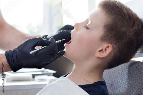 Ręce dentysty w jednorazowych czarnych gumowych rękawiczkach trzymają ekskawator dentystyczny. Chłopiec w wieku szkolnym siedzi na fotelu z otwartymi ustami. Lekarz ogląda zęby chłopca.