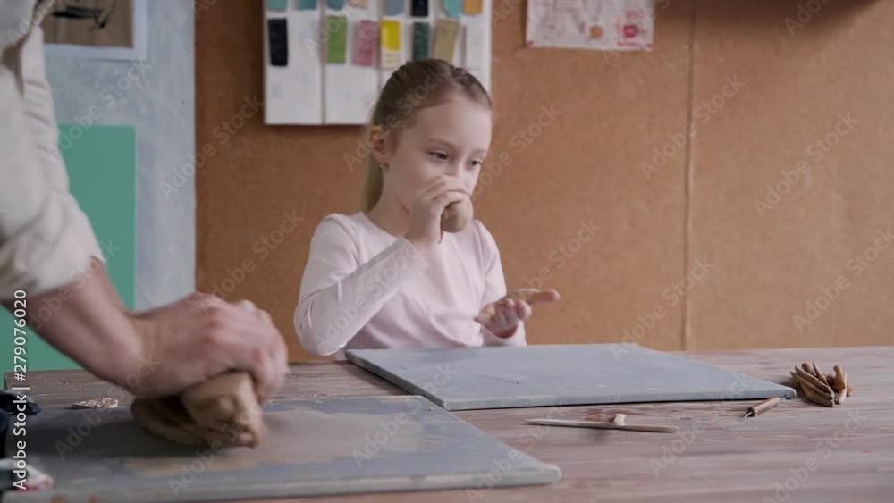 Adult craft man standing near little kid girl with calm face. Two person working with wet clay inside small studio or work space with natural light. They spending free time together and heaving fun Adult craft man standing near little kid girl with calm face. Two person working with wet clay inside small studio or work space with natural light. They spending free time together and heaving fun