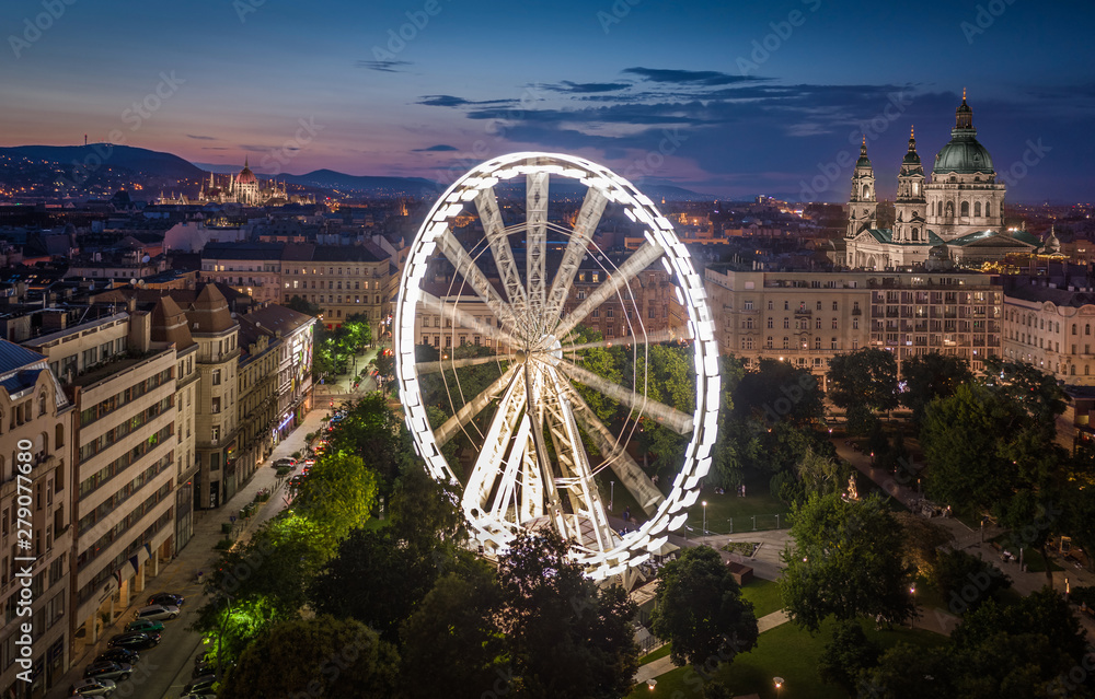 Fototapeta premium Budapeszt, Węgry - widok z lotu ptaka na plac Elżbiety o zmierzchu z oświetlonym diabelskim młynem, bazyliką św. Stefana i węgierski budynek parlamentu w tle. Letni wieczór w Budapeszcie