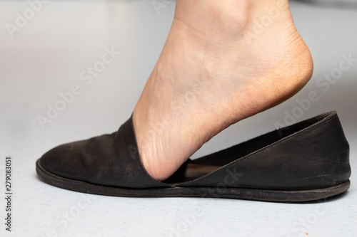 An extreme closeup and side view on the foot of a Caucasian lady putting footwear on. Standing on toes to slip black pump shoe over foot. Hardened skin near base of foot.
