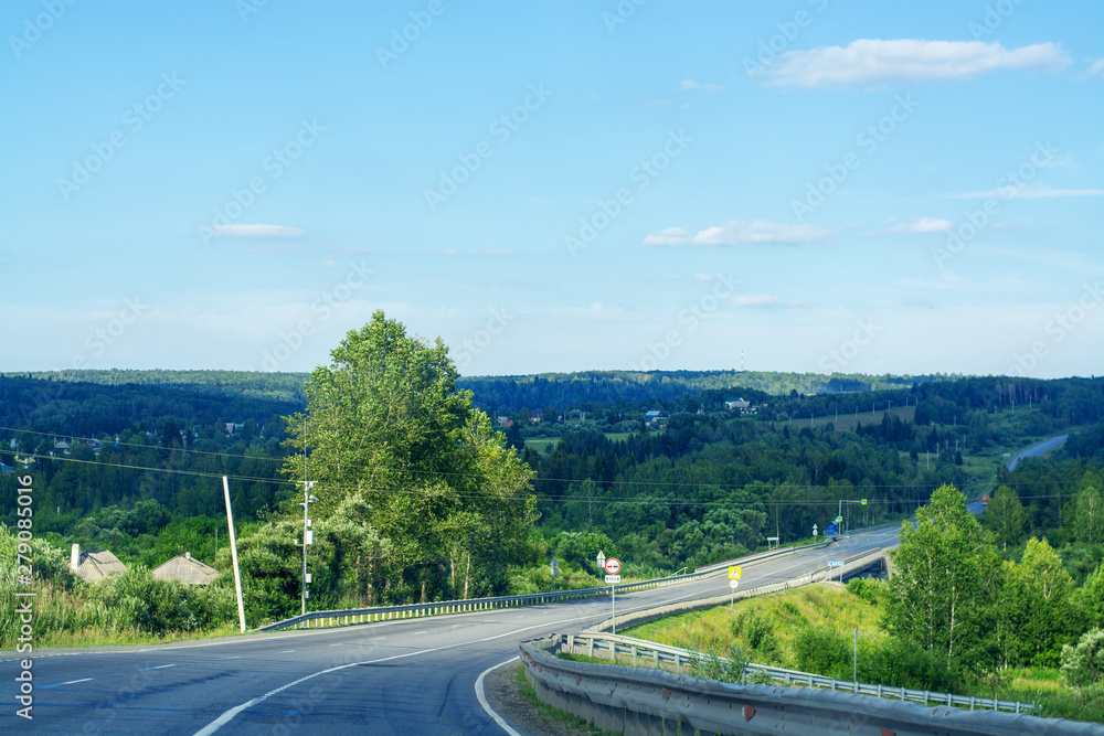 Asphalt road going through the forest on a summer sunny day. Around the green trees and Christmas trees. Clear blue sky. Journey by Siberia