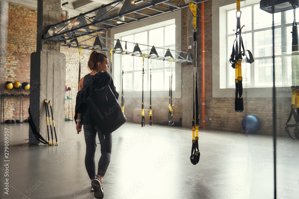 © Friends Stock - Morning workout. Back view of young athletic woman carrying her sport bag and looking aside while standing at empty gym.