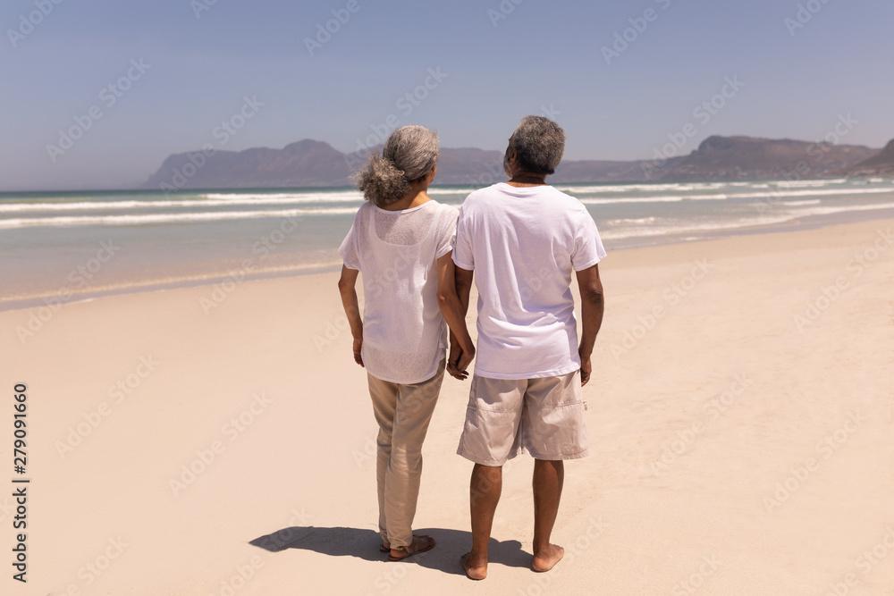 Senior couple holding hands and walking on beach with mountains in the background Senior couple holding hands and walking on beach with mountains in the background
