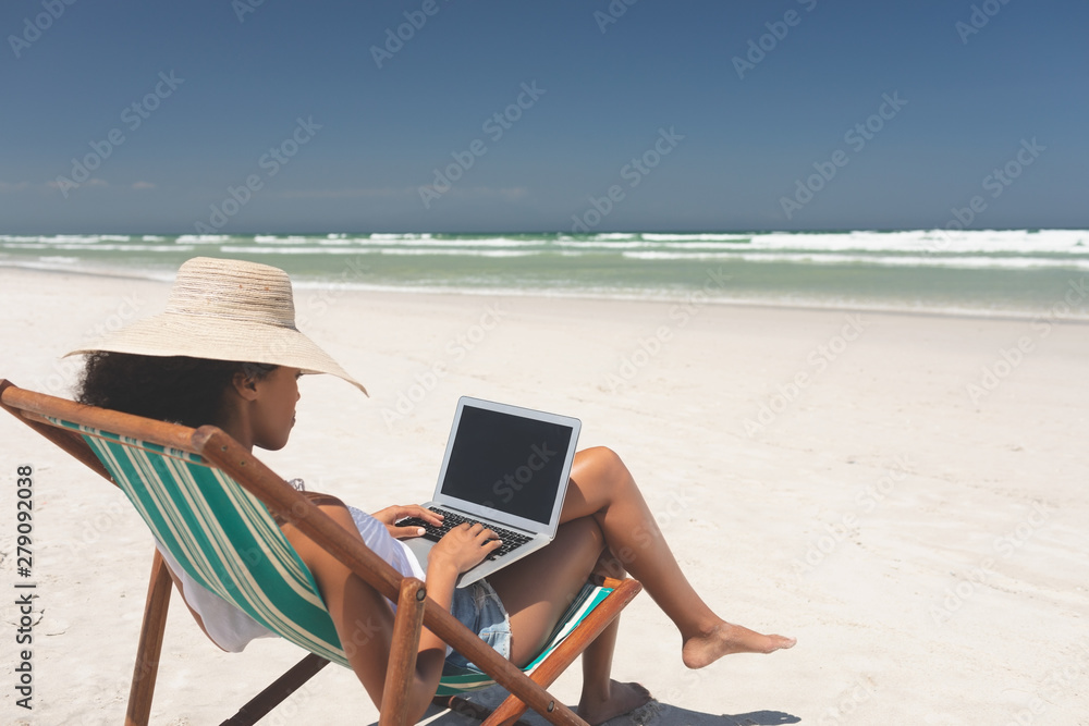 Young woman holding laptop while sitting on sun lounger at beach 