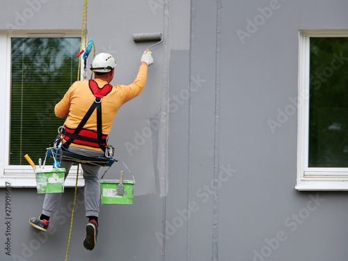 Worker hanging on rope and paints building wall with roller. Painter hanging on cable with paint buckets, industrial climber repairing house facade. Industrial alpinist and climbing. Rigging equipment