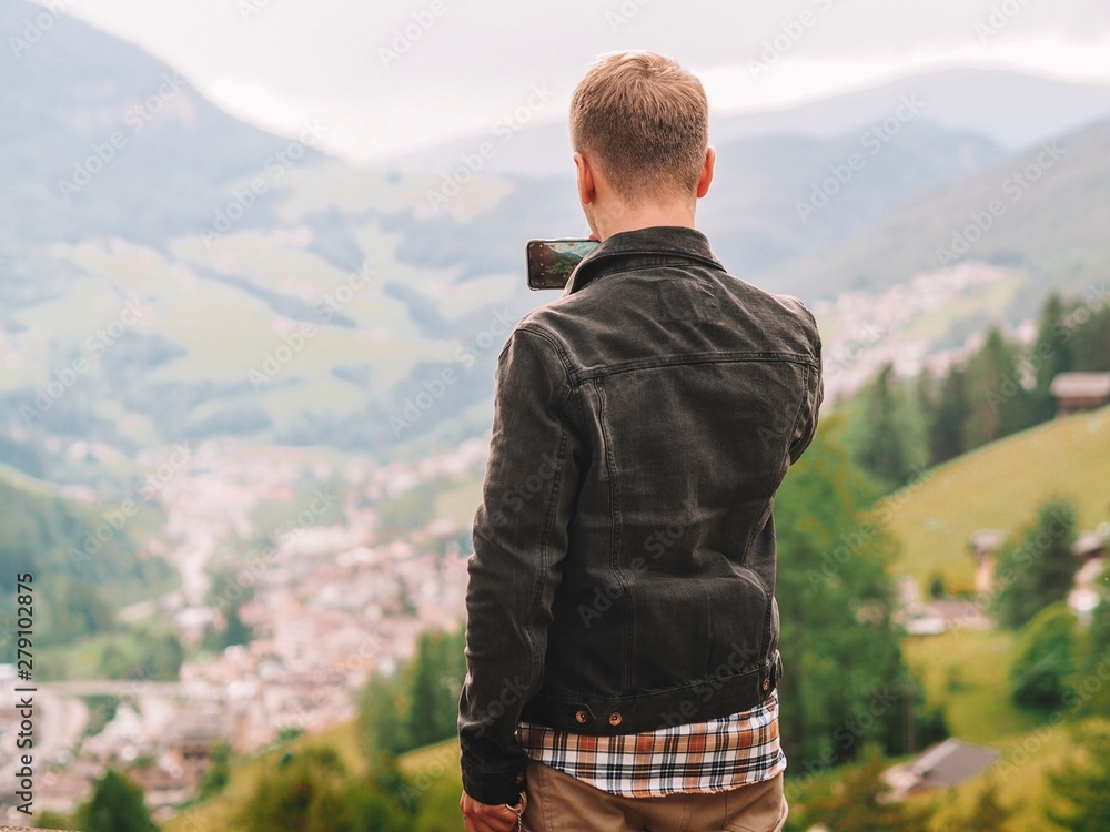 Naklejka premium A young man stands against the background of the dolomite mountains and photographs the landscape with his smartphone Mountain summer landscape in the Dolomites in Northern Italy.