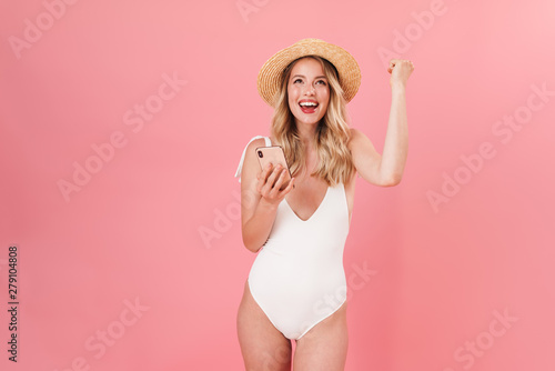 Pleased young cute blonde woman posing isolated over pink wall background in swimwear and hat using mobile phone make winner gesture.