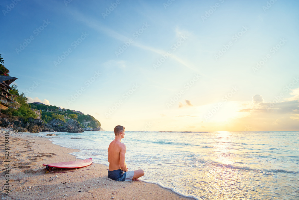 Surfing and meditation. Enjoying sunset.. Relaxed young man sitting on lotus position with surf board on the beach.