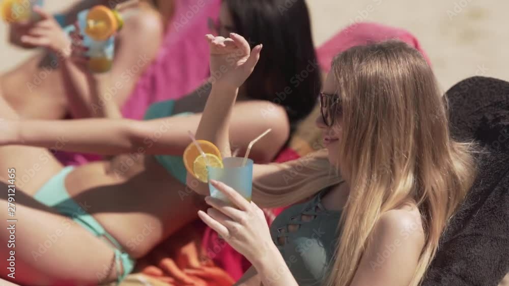 Three young beautiful models in a swimsuits are laying on a sunbeds on a beach, drinking a cocktails from a straws and dancing. Three young beautiful models in a swimsuits are laying on a sunbeds on a beach, drinking a cocktails from a straws and dancing.