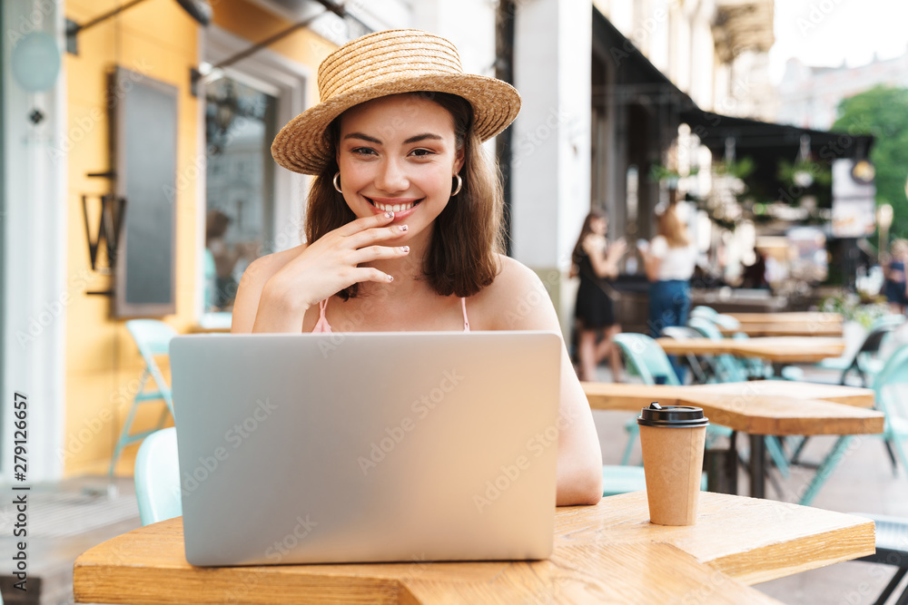 Image of satisfied brunette woman smiling and using laptop while sitting in street summer cafe
