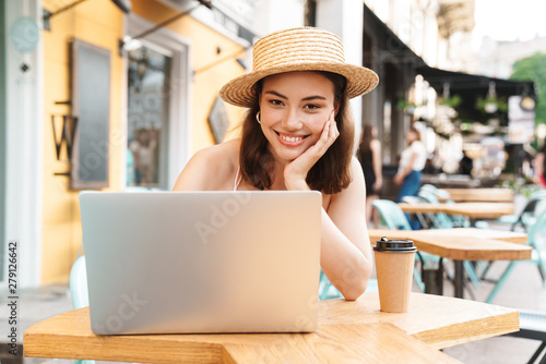 Image of joyful brunette woman smiling and using laptop while sitting in street summer cafe