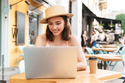 Image of pleased brunette woman smiling and using laptop while sitting in street summer cafe