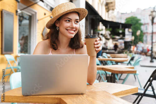 Image of elegant brunette woman smiling and using laptop while sitting in street summer cafe