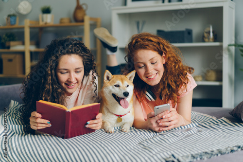 Girls friends are reading book and using smartphone lying on bed with cute shiba inu dog in cozy apartment. Modern lifestyle, friendship and animals concept.