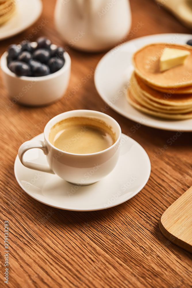 selective focus of cup of coffee with foam near plate with pancakes and bowl with strawberries selective focus of cup of coffee with foam near plate with pancakes and bowl with strawberries