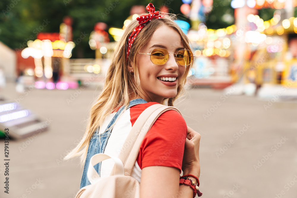 Image of pleased blonde woman smiling and walking in front of colorful carousel at amusement park - obrazy, fototapety, plakaty 