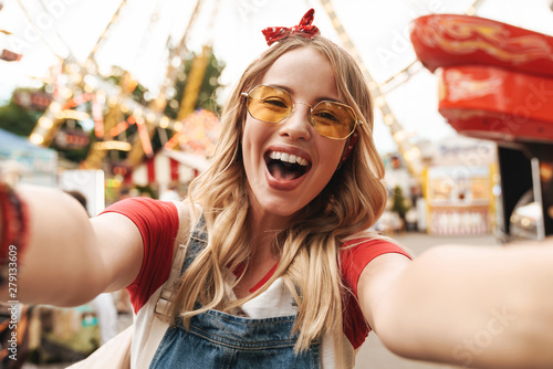 Image of cheerful blonde woman laughing and taking selfie photo at amusement park