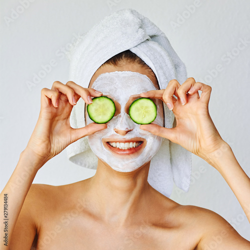 Beautiful young woman with facial mask on her face holding slices of cucumber...