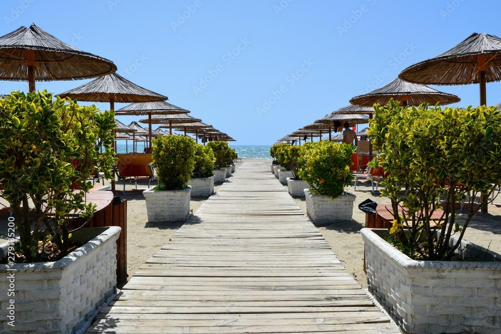 Obraz premium Wooden walkway leading to the beach and the coast between flower pots and beach umbrellas
