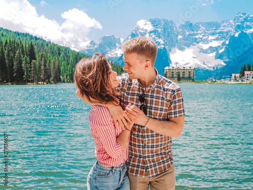 A girl with long hair in a striped shirt embraces a young man with a blond against the background of lake Mizurina in the Dolomites mountains, Italy. Amazing view of the lake. photo