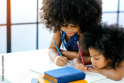 Vászonkép African girls as older and younger sister write or draw something on white paper near the book in front of glass windows with day light