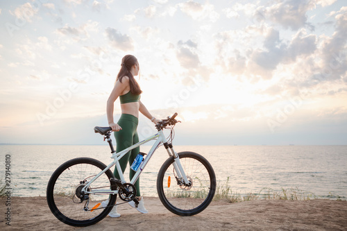 Beautiful young brunette woman resting after bicycle ride at sunrise. Model listening to music with wireless earbuds in the morning.